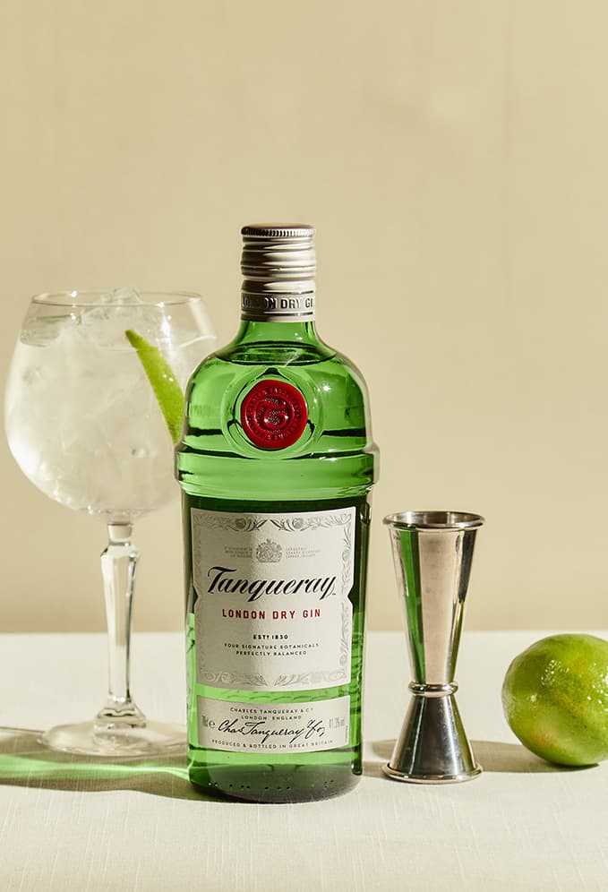 A Gin & Tonic cocktail made with Tanqueray London Dry Gin garnished with two lime slices. A bottle of Tanqueray London Dry gin sits in the background of the image.