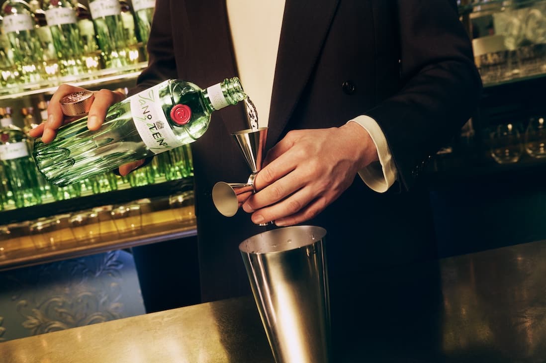 A hand pouring Tanqueray No. Ten into a 4 way jigger in front of a cocktail shaker.