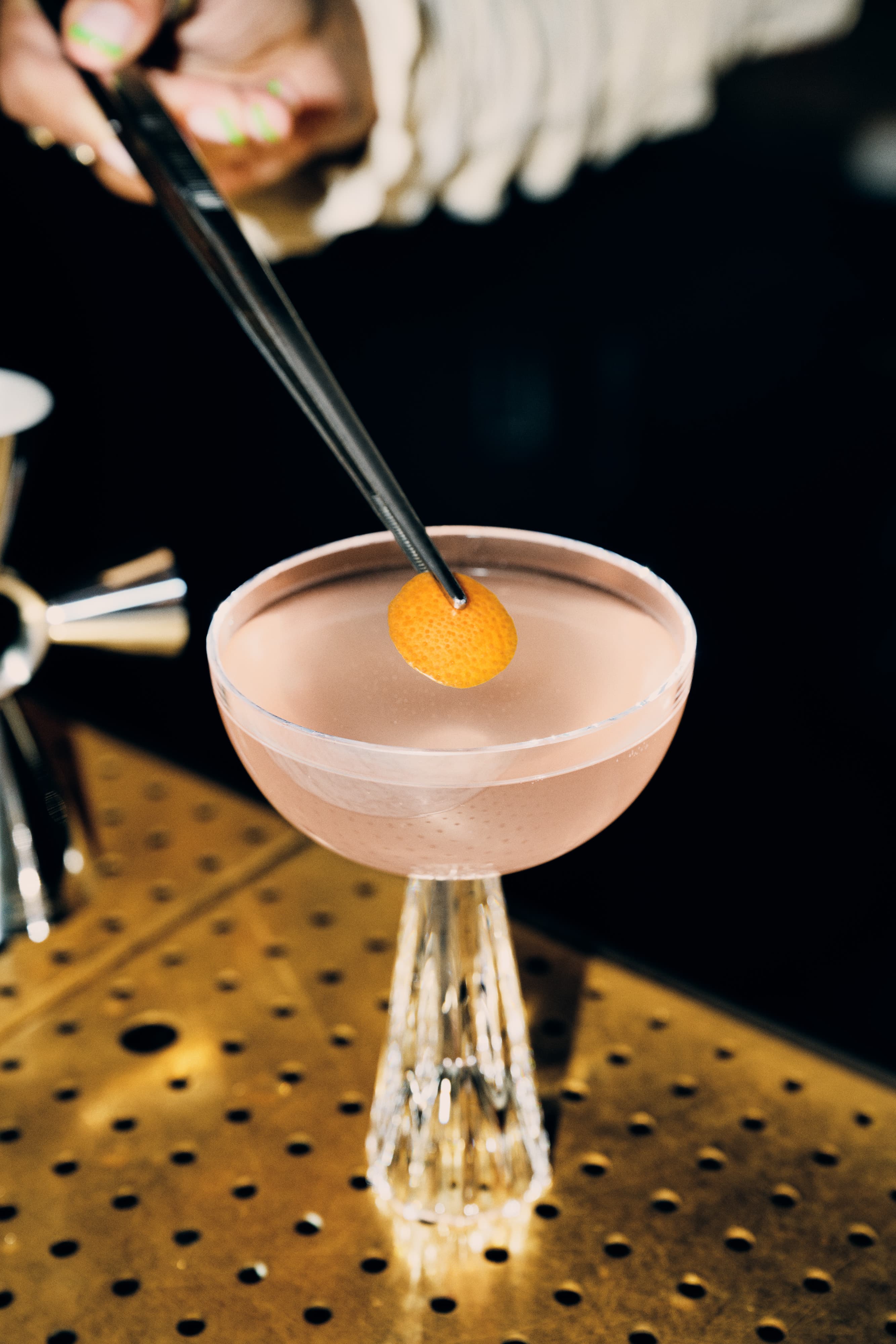 Tongs placing a grapefruit peel on top of a Tiny Ten cocktail
