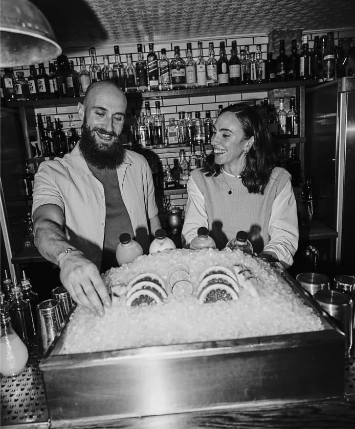 Keila & Yoann standing behind an ice box of garnishes, including grapefruit