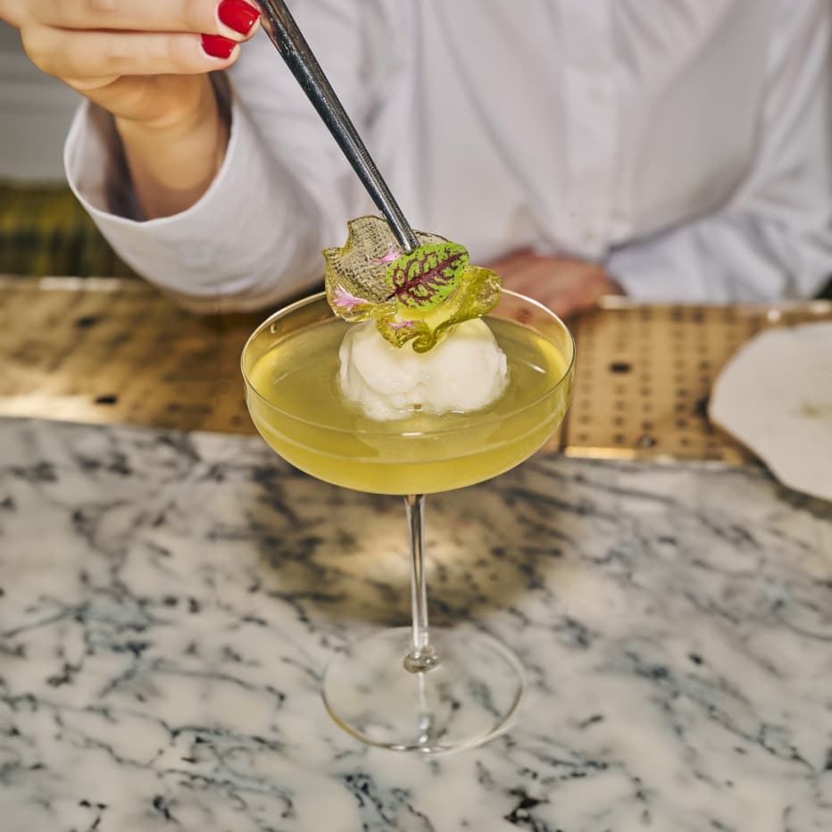A cocktail glass sits on a marble counter, close behind we see the arm and shoulders person holding some tweezers to place the final flourish of a small green leaf onto the cocktail.