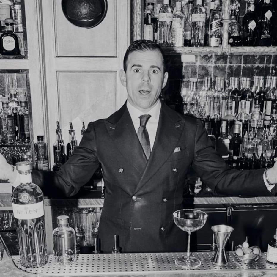 A black and white image of a man standing behind a bar. On the bar in front of him is an array of cocktail making equipment and a bottle of Tanqueray No. TEN.